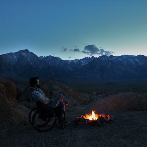 ryan clarkson seated in a wheelchair in the wilderness at dusk by a campfire.