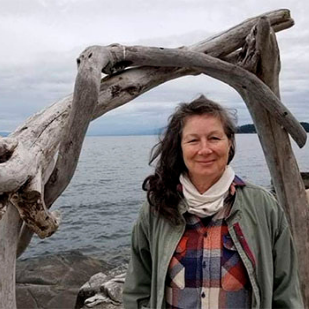Bobbie Seale-Cobiskey stands in under a wooden structure in front of the ocean. She is wearing a plaid shirt and a green jacket.