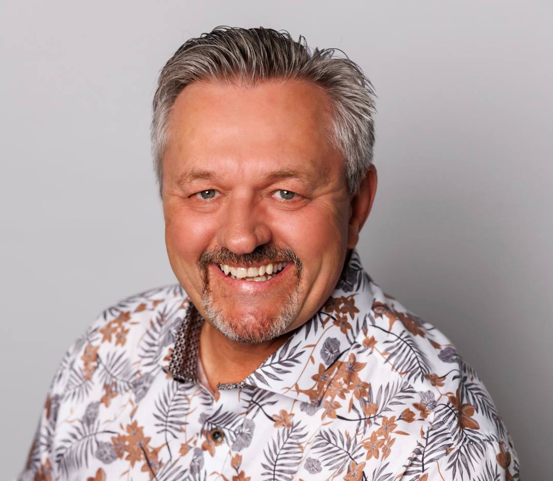 Headshot of Nate Toevs smiling, wearing a white and gold floral button-up shirt.